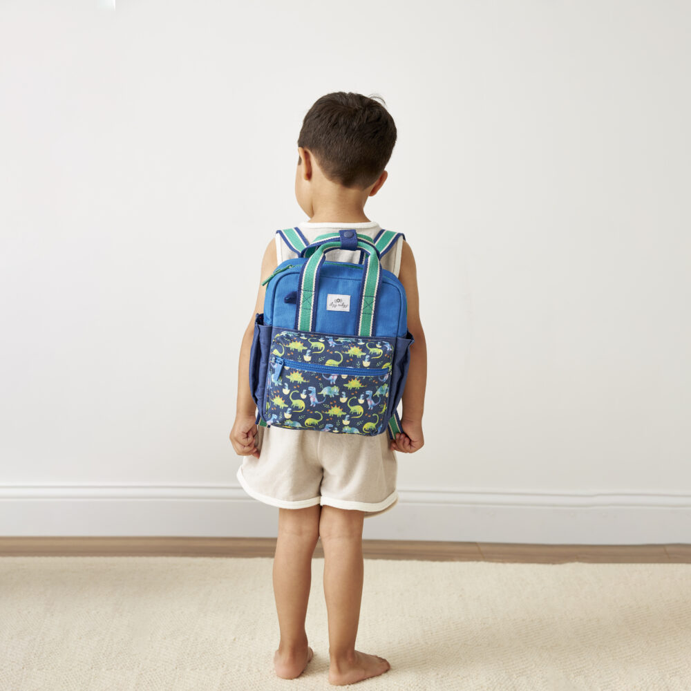 A young child with short hair stands facing away, wearing a white tank top, beige shorts, and a blue backpack with a floral pattern. The background features a plain wall and a light carpet. The child is accessorized with an Itzy Ritzy Sweetie Strap Plus Bear Silicone Pacifier Clip.