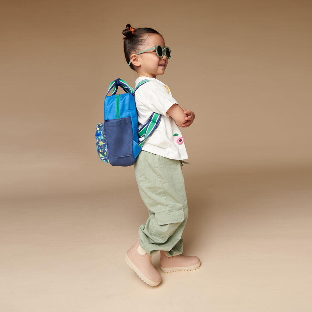 Young child with a top knot hairstyle, sunglasses, and a backpack, wearing a white shirt, green pants, beige boots, and the Itzy Ritzy Sweetie Strap Plus Bear Silicone Pacifier Clip, standing against a plain background.