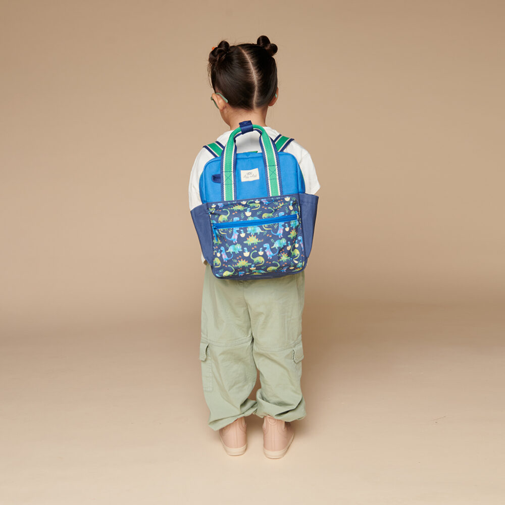 A young child facing away, standing against a beige background, is wearing a blue backpack with a dinosaur pattern, green pants, a white shirt, pink shoes, and sporting an Itzy Ritzy Sweetie Strap Plus Bear Silicone Pacifier Clip.