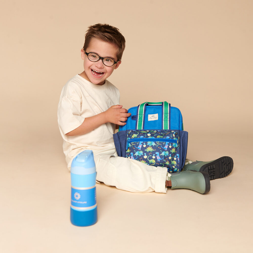 A child with short hair and glasses sits on the floor, smiling and holding an Itzy Ritzy Sweetie Strap Plus Bear Silicone Pacifier Clip. A blue water bottle is placed in the foreground.