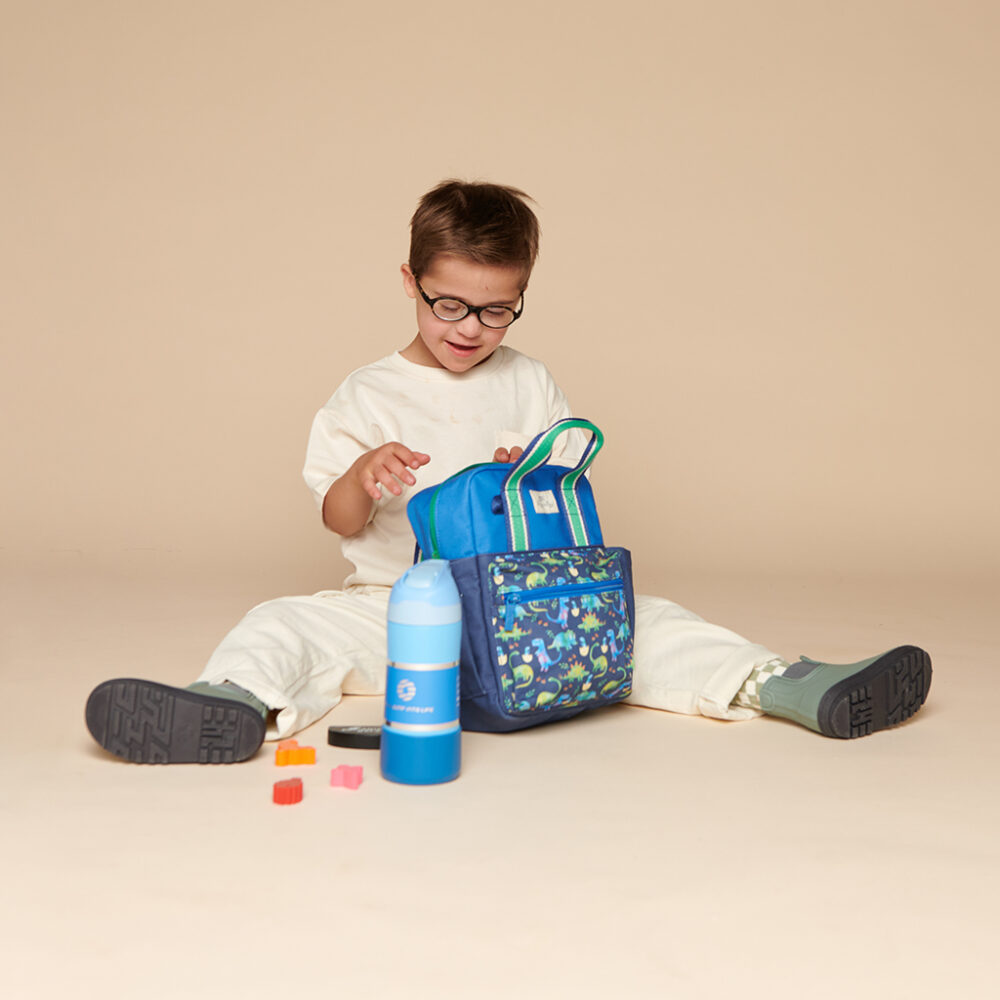 A young child wearing glasses sits on the floor with legs spread, looking into a blue backpack. A water bottle and some small colorful blocks are placed in front, along with an Itzy Ritzy Sweetie Strap Plus Bear Silicone Pacifier Clip.