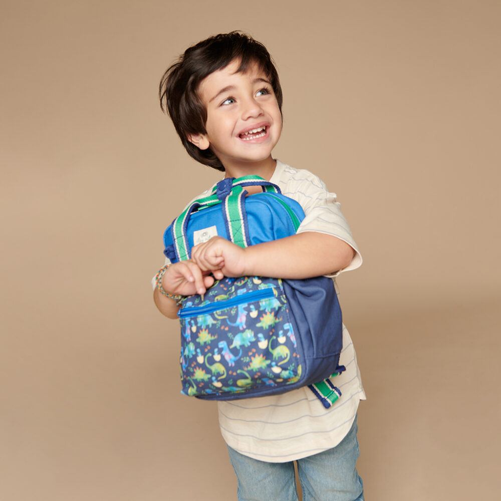 A young child smiling while holding an Itzy Ritzy Sweetie Strap Plus Bear Silicone Pacifier Clip, standing in front of a plain beige background.