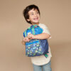 A young child smiling while holding an Itzy Ritzy Sweetie Strap Plus Bear Silicone Pacifier Clip, standing in front of a plain beige background.