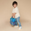 Young child wearing a striped shirt and jeans holds an Itzy Ritzy Sweetie Strap Plus Bear Silicone Pacifier Clip, standing against a plain beige background.