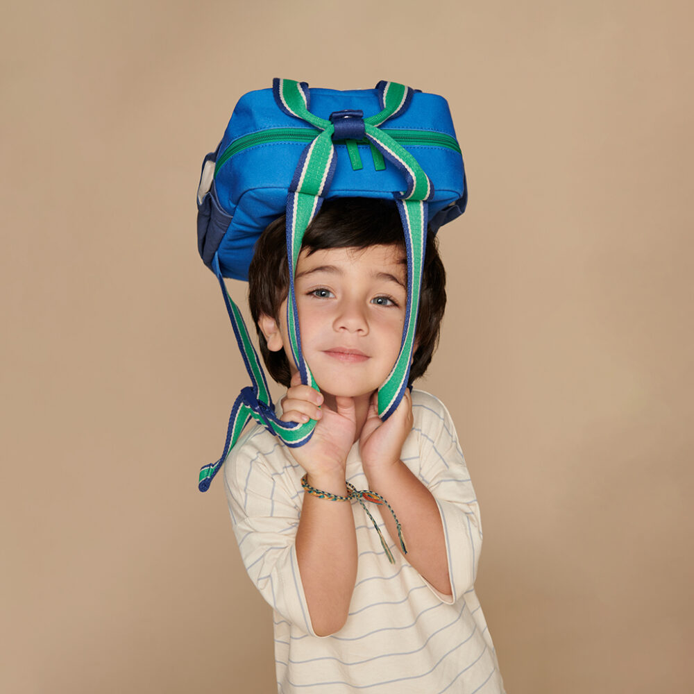 A young boy with dark hair is wearing the Itzy Ritzy Sweetie Strap Plus Bear Silicone Pacifier Clip as a blue backpack on his head, with his hands holding the straps under his chin. He is dressed in a white and light blue striped shirt.