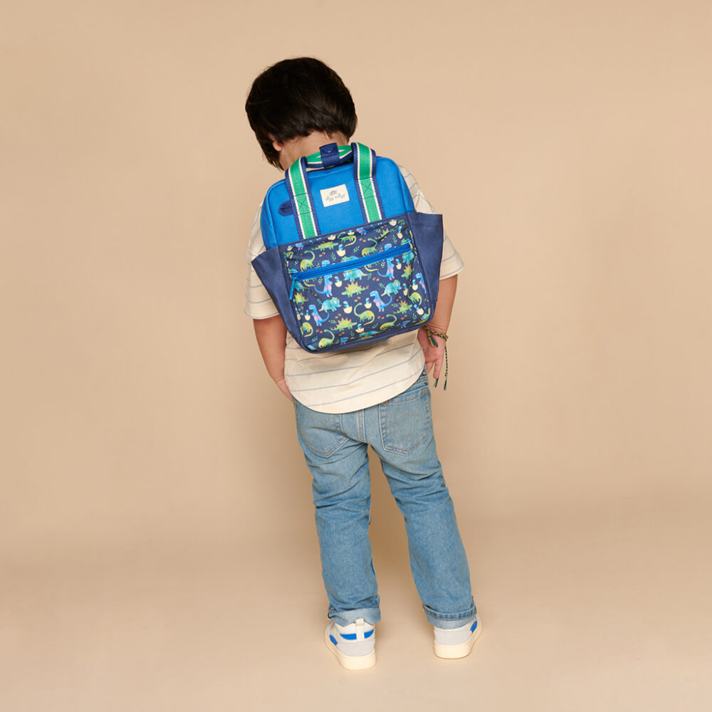 A child wearing jeans and a striped shirt, with an Itzy Ritzy Sweetie Strap Plus Bear Silicone Pacifier Clip attached to their blue and green patterned backpack, standing with their back to the camera against a plain beige background.