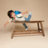 A child with an Itzy Ritzy Sweetie Strap Plus Bear Silicone Pacifier Clip attached to their backpack is playfully lying across a wooden table, with one arm and one leg extended in the air.