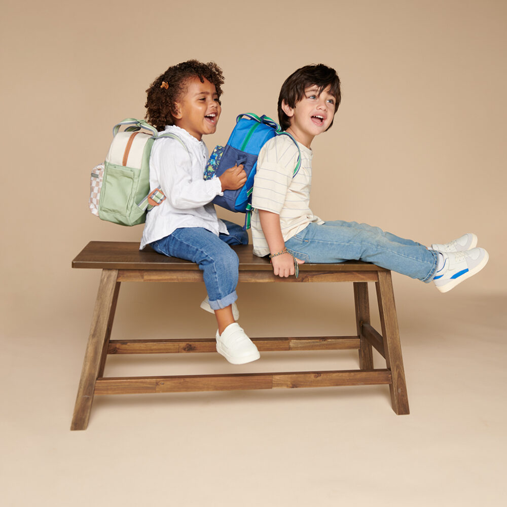 Two children with backpacks sit and laugh on a wooden bench against a plain background. One child, wearing a white top, is using an Itzy Ritzy Sweetie Strap Plus Bear Silicone Pacifier Clip, while the other child wears a striped shirt.