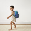 A young boy smiles while walking barefoot, carrying an Itzy Ritzy Sweetie Strap Plus Bear Silicone Pacifier Clip, in a light-colored room with a beige rug.