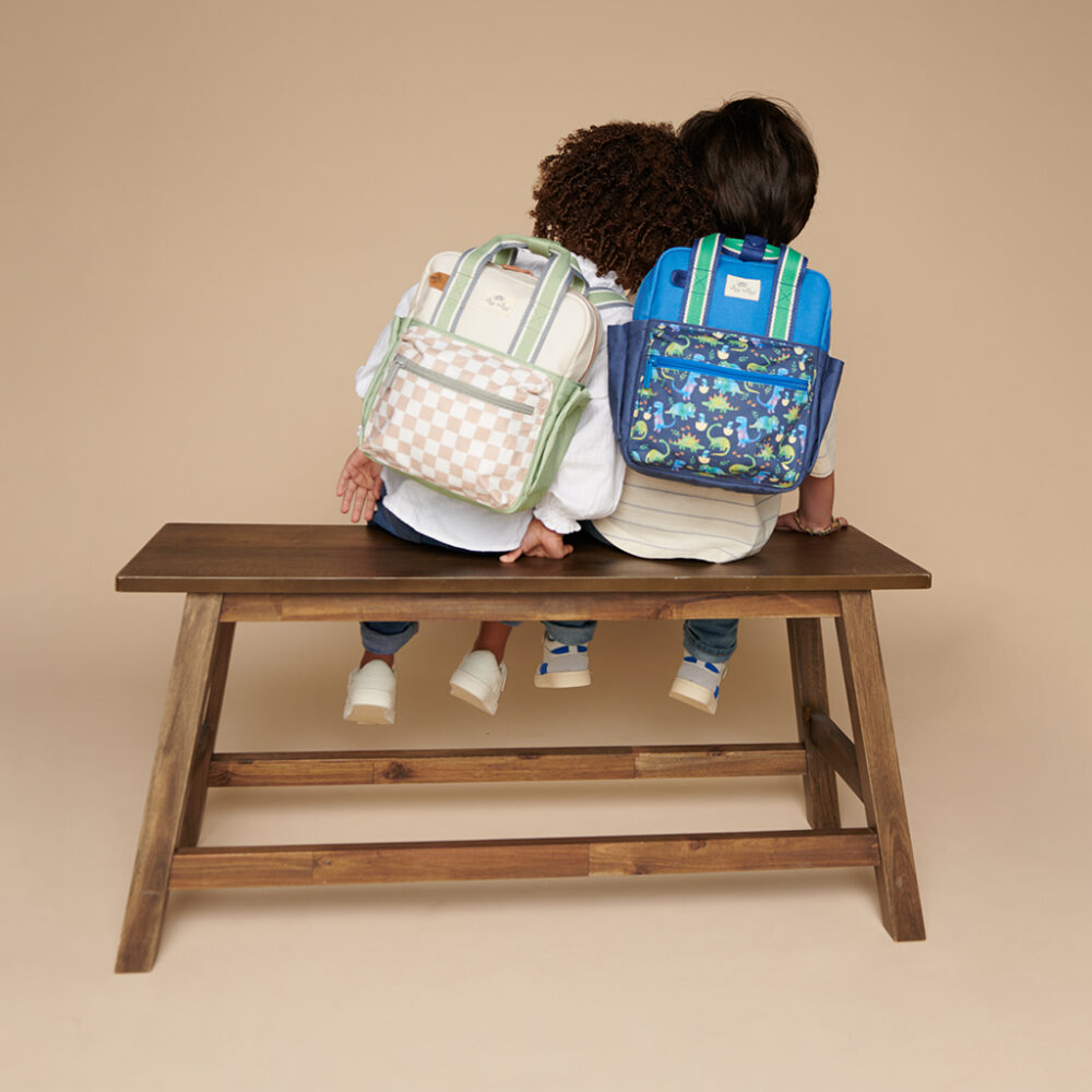 Two children wearing colorful backpacks sit side by side on a wooden bench, both looking away from the camera, with one child holding an Itzy Ritzy Sweetie Strap Plus Bear Silicone Pacifier Clip.