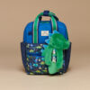 A blue and green children's backpack featuring a dinosaur design, with an attached plush toy similar to the Itzy Ritzy Sweetie Strap Plus Bear Silicone Pacifier Clip.