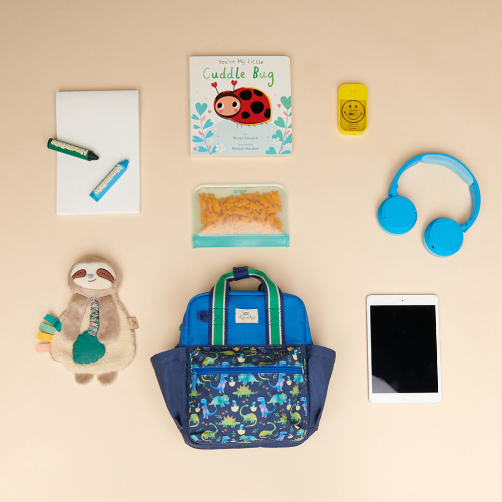 A neatly arranged assortment featuring an Itzy Ritzy Sweetie Strap Plus Bear Silicone Pacifier Clip, a stuffed sloth, a book titled "You're My Little Cuddle Bug," headphones, tablet, notebook with pencil, lunchbox with snacks, smiley face toy, and child's backpack.