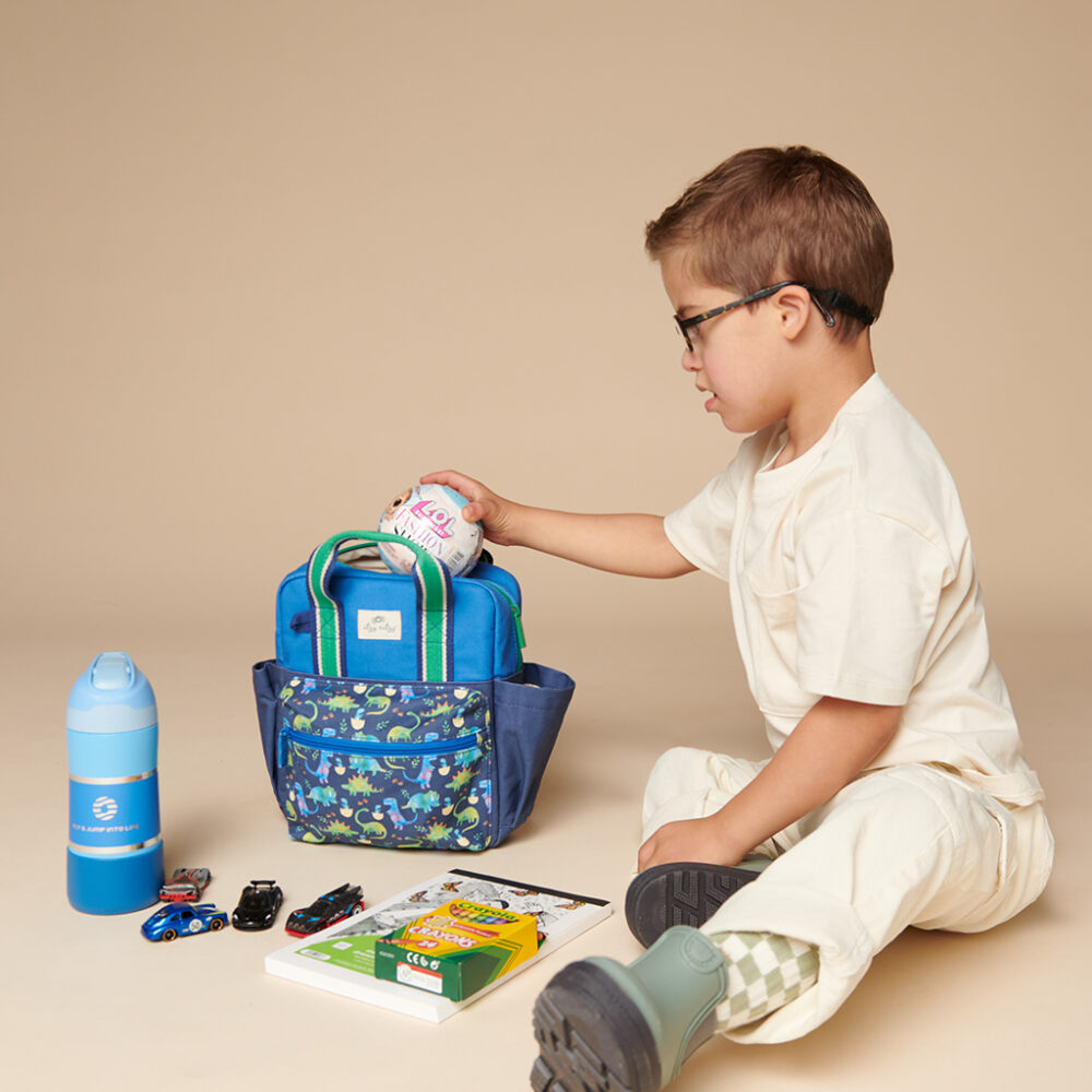 A child wearing glasses and a light-colored outfit places an item in a blue and green backpack. Nearby are small toy cars, a water bottle, books, the Itzy Ritzy Sweetie Strap Plus Bear Silicone Pacifier Clip, and other school supplies on the floor.