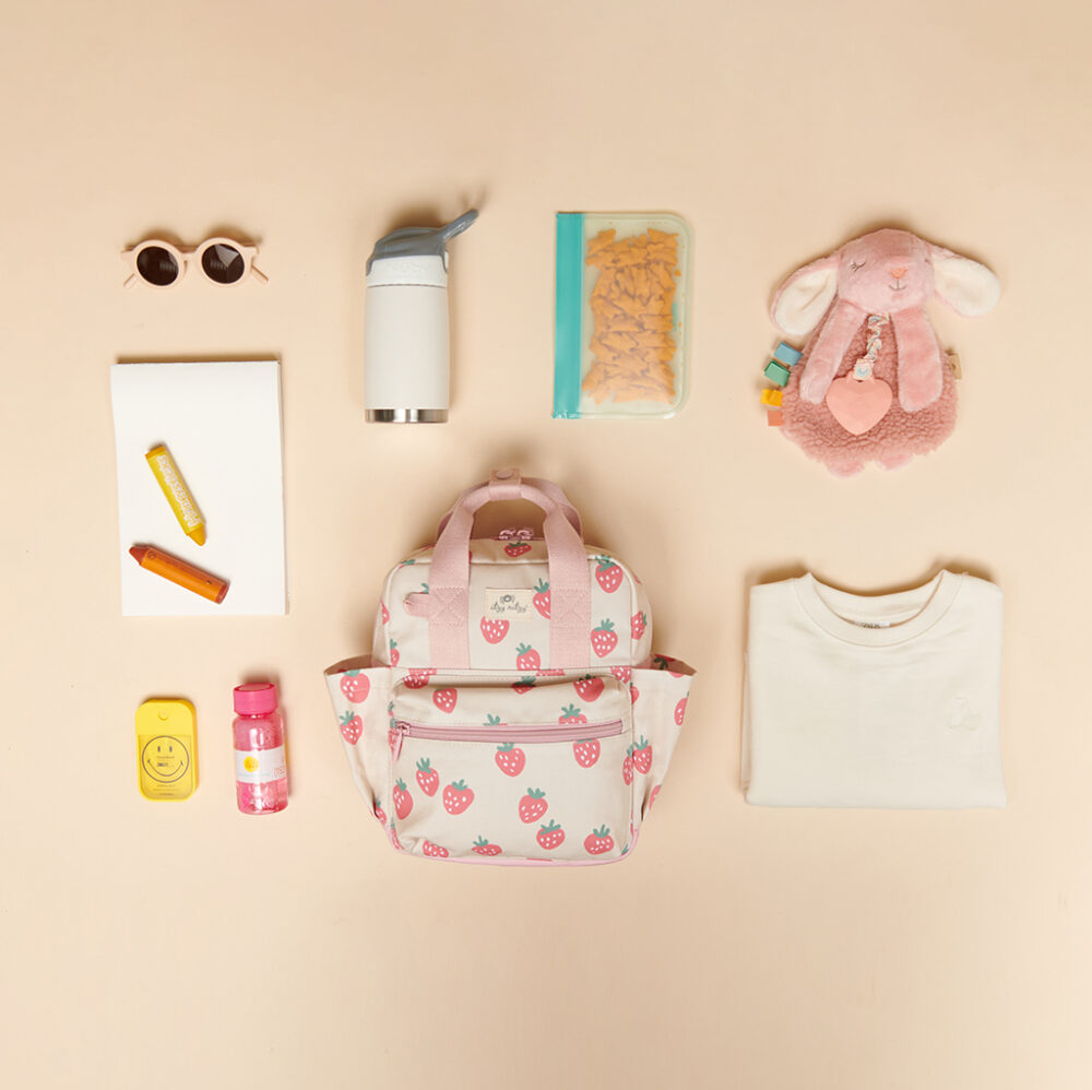 A small backpack with a strawberry design is surrounded by sunglasses, a thermos, snacks in a bag, a toy bunny, a notebook, a crayon, lip balm, a snack container, and an Itzy Ritzy Sweetie Strap Plus Bear Silicone Pacifier Clip.