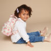 A young child with curly hair, wearing a white shirt, jeans, and beige boots, sits on the floor with a strawberry-patterned backpack and a purple bow in their hair, accessorized with an Itzy Ritzy Sweetie Strap Plus Bear Silicone Pacifier Clip.