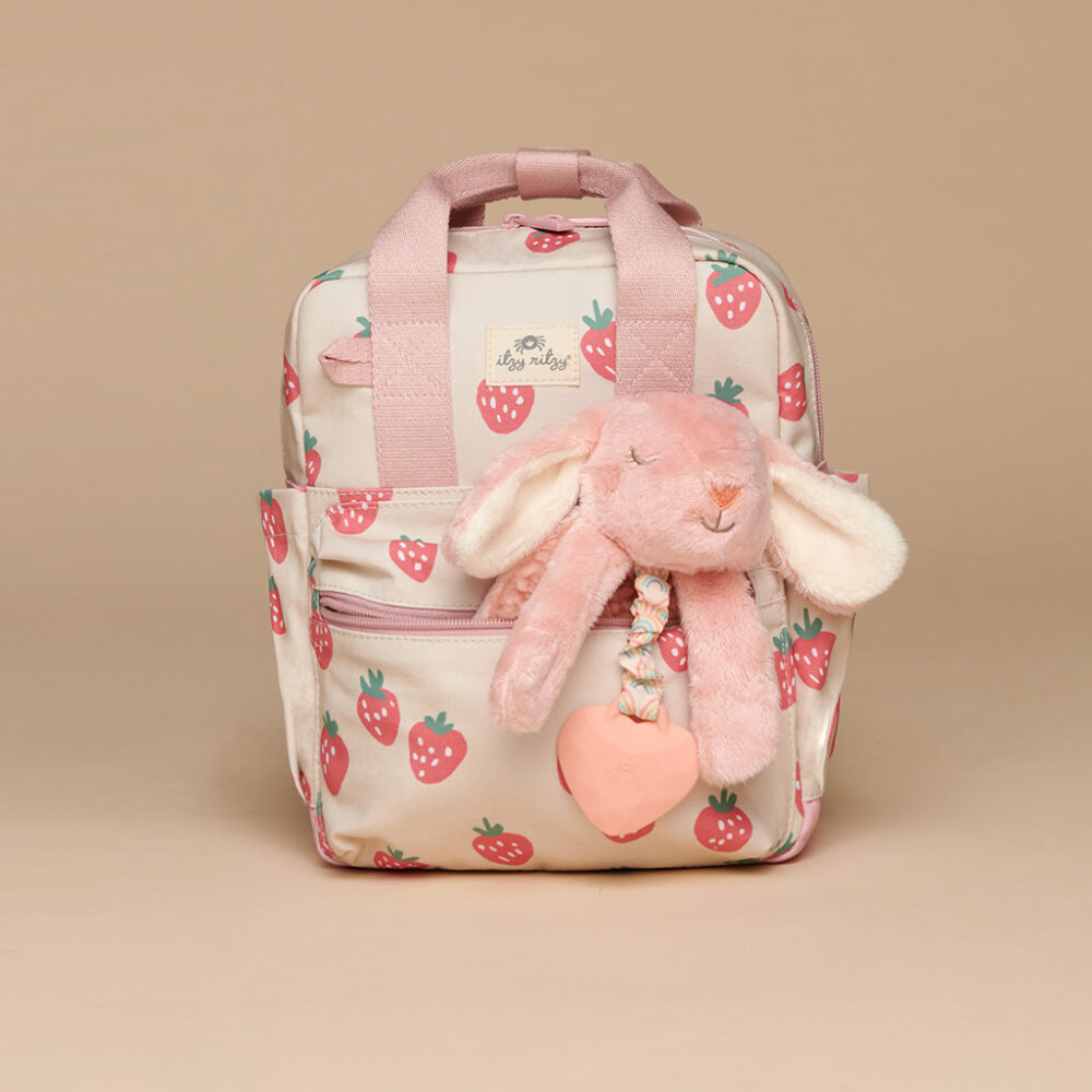 A small backpack with pink handles and strawberry patterns, featuring the Itzy Ritzy Sweetie Strap Plus Bear Silicone Pacifier Clip attached to the front pocket.