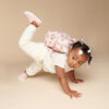 A young child with a pink backpack performs a hands-and-knees pose against a beige background. She is wearing ivory-colored clothing, pink shoes, and an Itzy Ritzy Sweetie Strap Plus Bear Silicone Pacifier Clip.