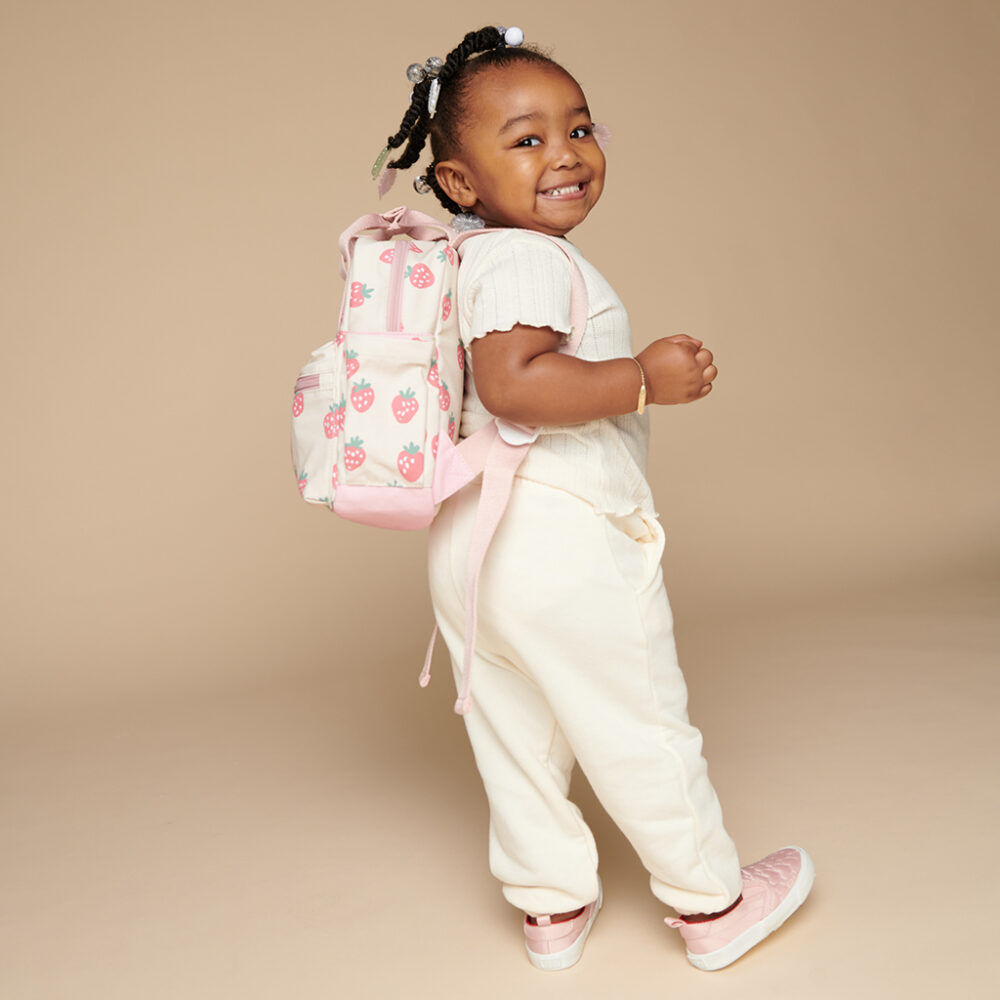 A young child with braided hair smiles and poses sideways, wearing a white outfit, pink shoes, and carrying an adorable backpack with a strawberry pattern, accessorized perfectly with the Itzy Ritzy Sweetie Strap Plus Bear Silicone Pacifier Clip.