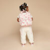 A toddler wearing cream-colored attire stands facing away on a neutral background, showcasing a pink strawberry-patterned backpack and adorning their hairstyle with white and pink hair clips, along with an Itzy Ritzy Sweetie Strap Plus Bear Silicone Pacifier Clip.