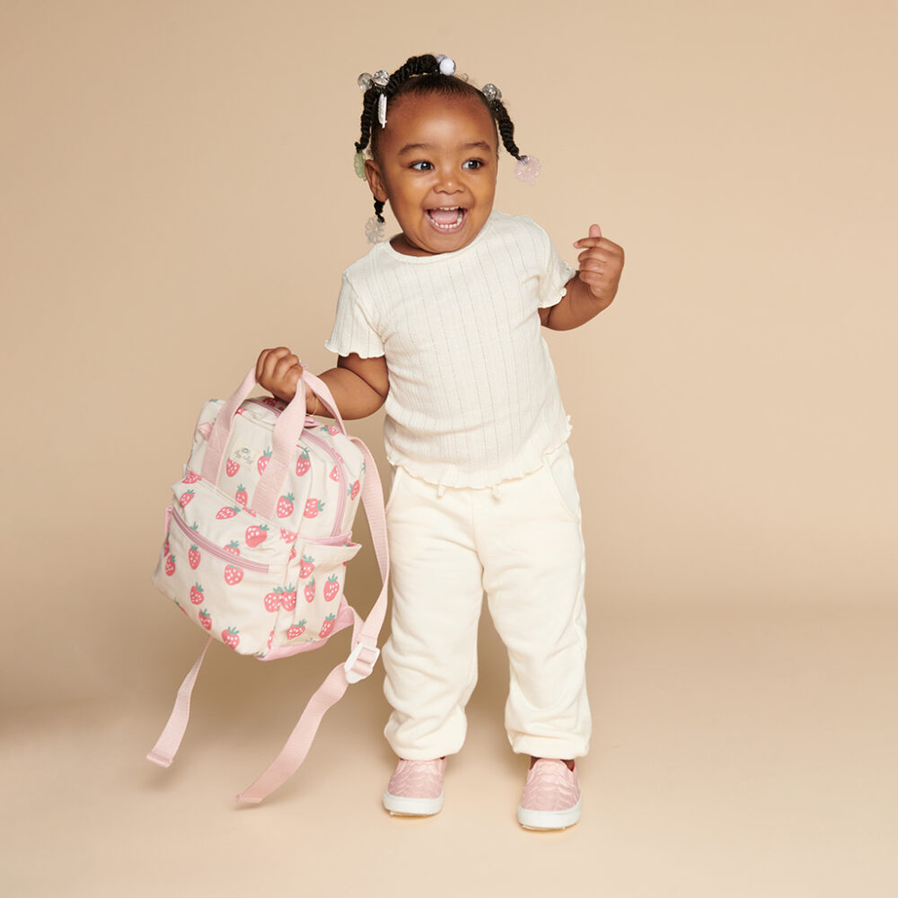 A smiling toddler stands against a beige background, holding an Itzy Ritzy Sweetie Strap Plus Bear Silicone Pacifier Clip in one hand. The child wears a white shirt and pants, with pink shoes.