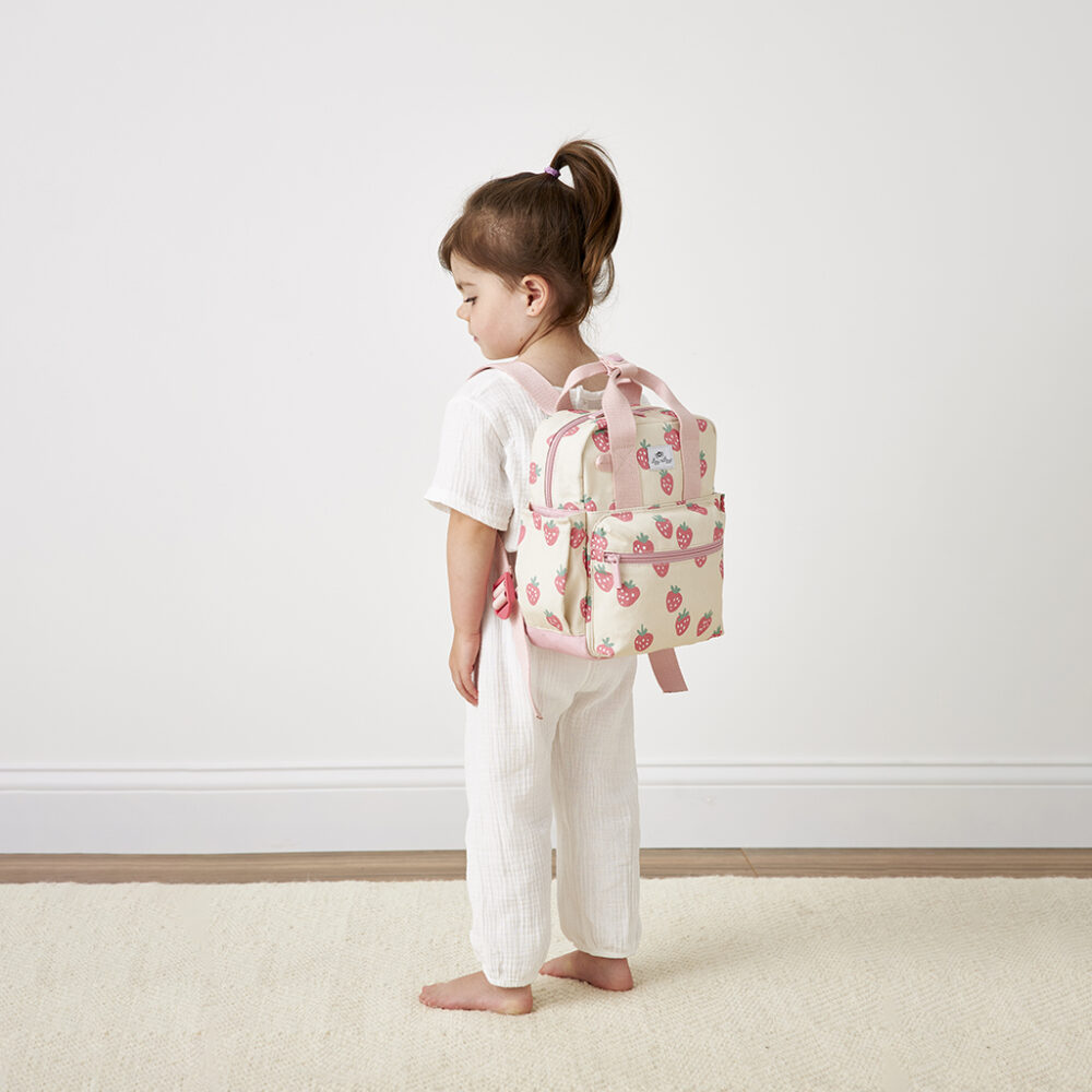 A young child with light brown hair stands in profile, clad in an all-white outfit and carrying a pink strawberry-patterned backpack. She is barefoot on a light-colored rug against a white wall, with the Itzy Ritzy Sweetie Strap Plus Bear Silicone Pacifier Clip clipped to her attire.