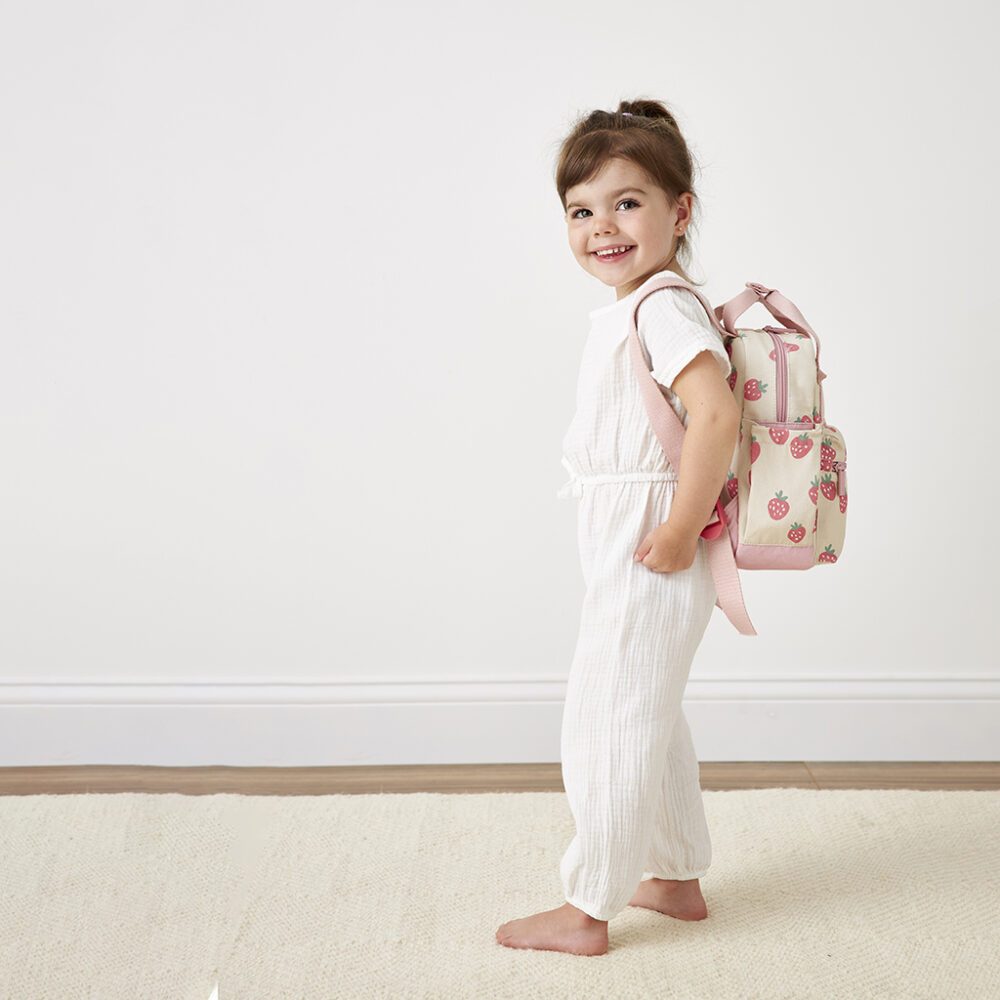 A smiling young girl in white clothes stands sideways, holding an Itzy Ritzy Sweetie Strap Plus Bear Silicone Pacifier Clip over her shoulder against a white background.