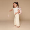 A young girl smiling while wearing a tiara, white shirt, cream pants, and ballet shoes holds a wand with an Itzy Ritzy Sweetie Strap Plus Bear Silicone Pacifier Clip visible on her floral pink backpack against a beige background.