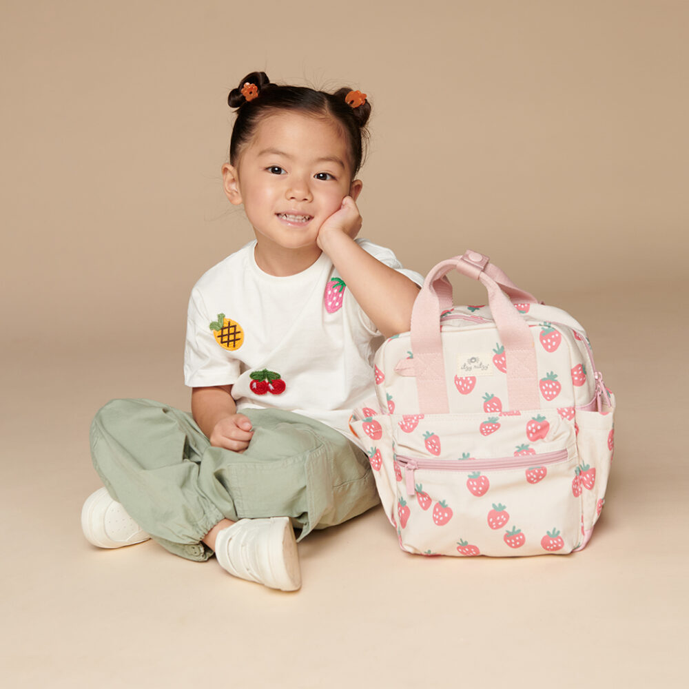 A young child sits on the floor with a hand on their cheek, wearing a white shirt with fruit graphics, green pants, and white shoes, next to an Itzy Ritzy Sweetie Strap Plus Bear Silicone Pacifier Clip in pink featuring strawberry prints.