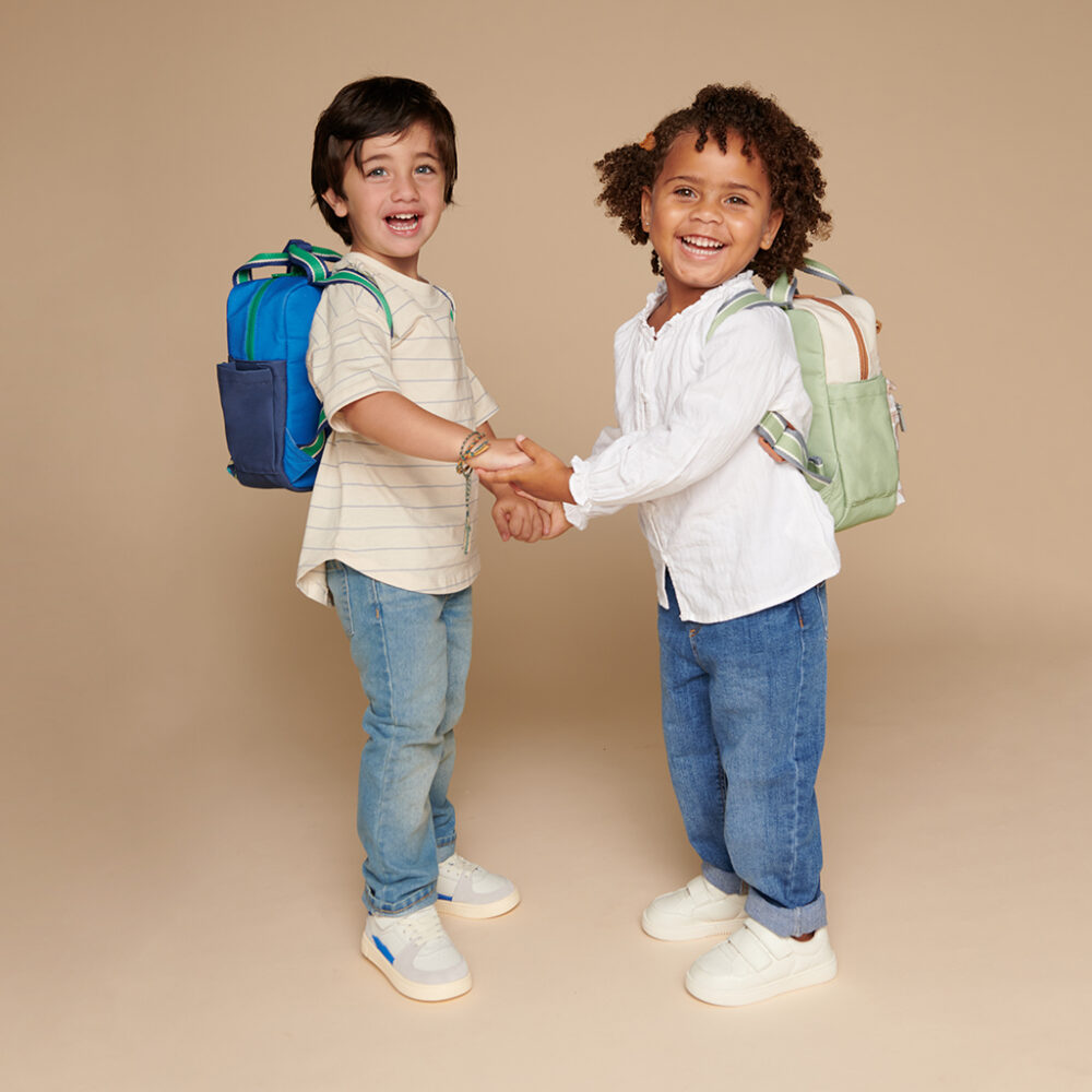 Two young children, each wearing an Itzy Ritzy Sweetie Strap Plus Bear Silicone Pacifier Clip on their backpacks, smile and hold hands while standing against a neutral background.