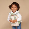 A young child with curly hair smiles while holding an Itzy Ritzy Sweetie Strap Plus Bear Silicone Pacifier Clip. The child is wearing a white long-sleeve shirt and blue jeans, set against a plain beige background.