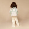 A child with long hair, facing away, stands against a beige background wearing a cream outfit and an Itzy Ritzy Sweetie Strap Plus Bear Silicone Pacifier Clip on their checked backpack with green straps.