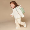 A young child with curly hair wears a beige sweater, light pants, and white shoes, carrying a light green backpack and an Itzy Ritzy Sweetie Strap Plus Bear Silicone Pacifier Clip against a neutral background.