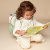 A young child with curly hair, wearing a white sweater and light-colored pants, sits on the floor reading a book titled "Hello Bunny" while sporting an Itzy Ritzy Sweetie Strap Plus Bear Silicone Pacifier Clip.