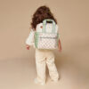 A child with long, curly hair stands facing away, wearing a Itzy Ritzy Sweetie Strap Plus Bear Silicone Pacifier Clip, featuring a large backpack with a checkered front pocket in cream and green colors. The child is dressed in light-colored casual clothing against a plain background.