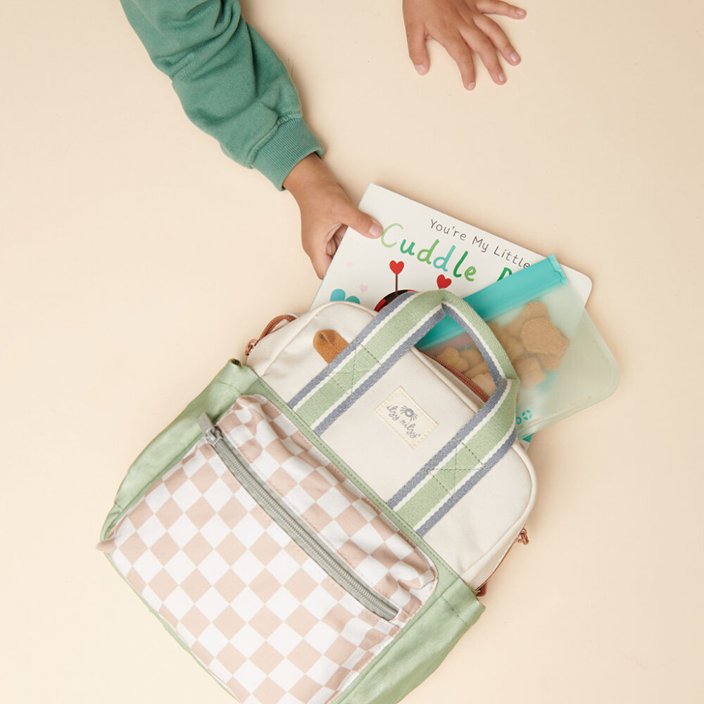 A child reaches for a book from a backpack containing a book titled "Cuddle Bug" and an Itzy Ritzy Sweetie Strap Plus Bear Silicone Pacifier Clip on a light-colored surface.