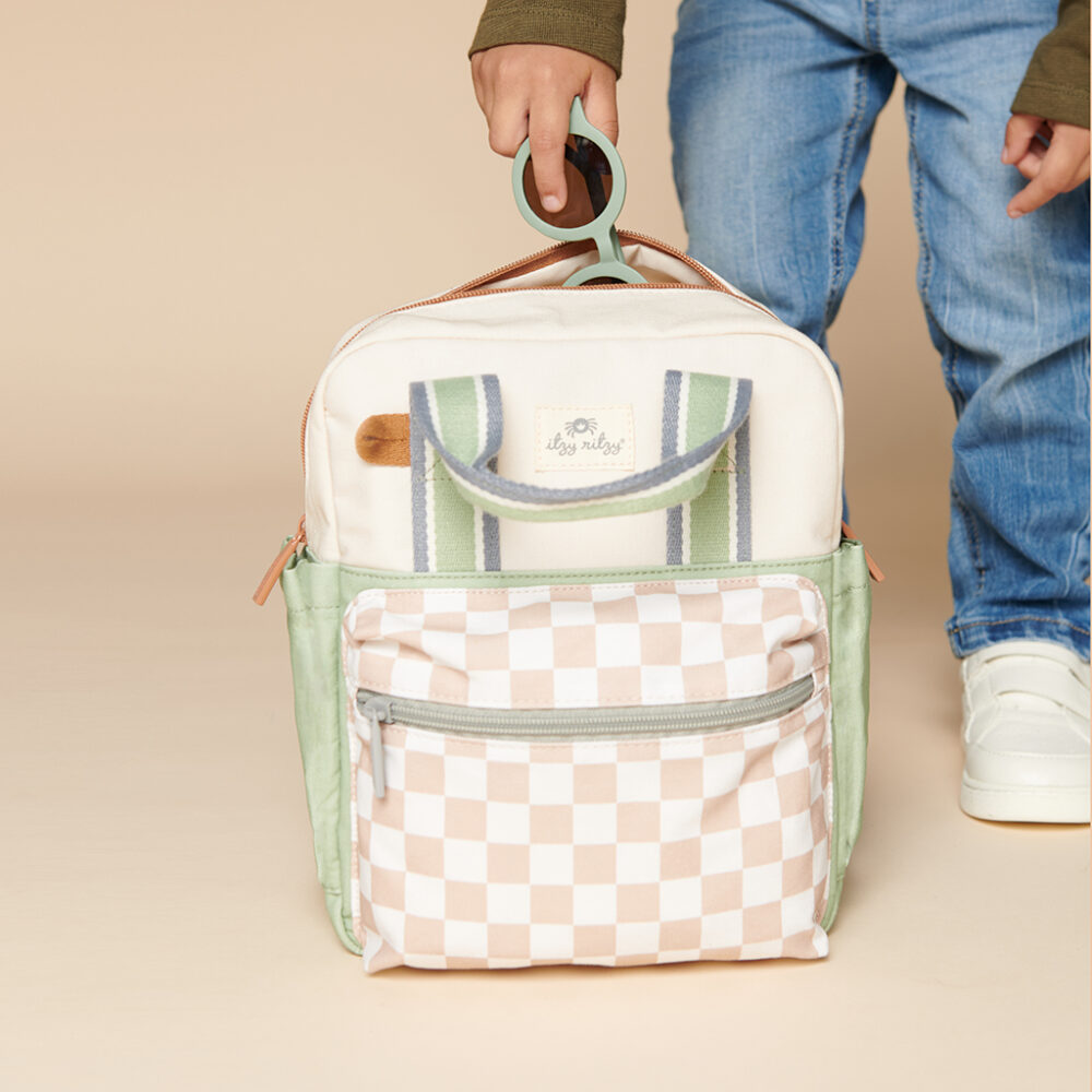 A child holds an Itzy Ritzy Sweetie Strap Plus Bear Silicone Pacifier Clip, featuring a checkered front pocket, green side accents, and striped handles. The background is beige.