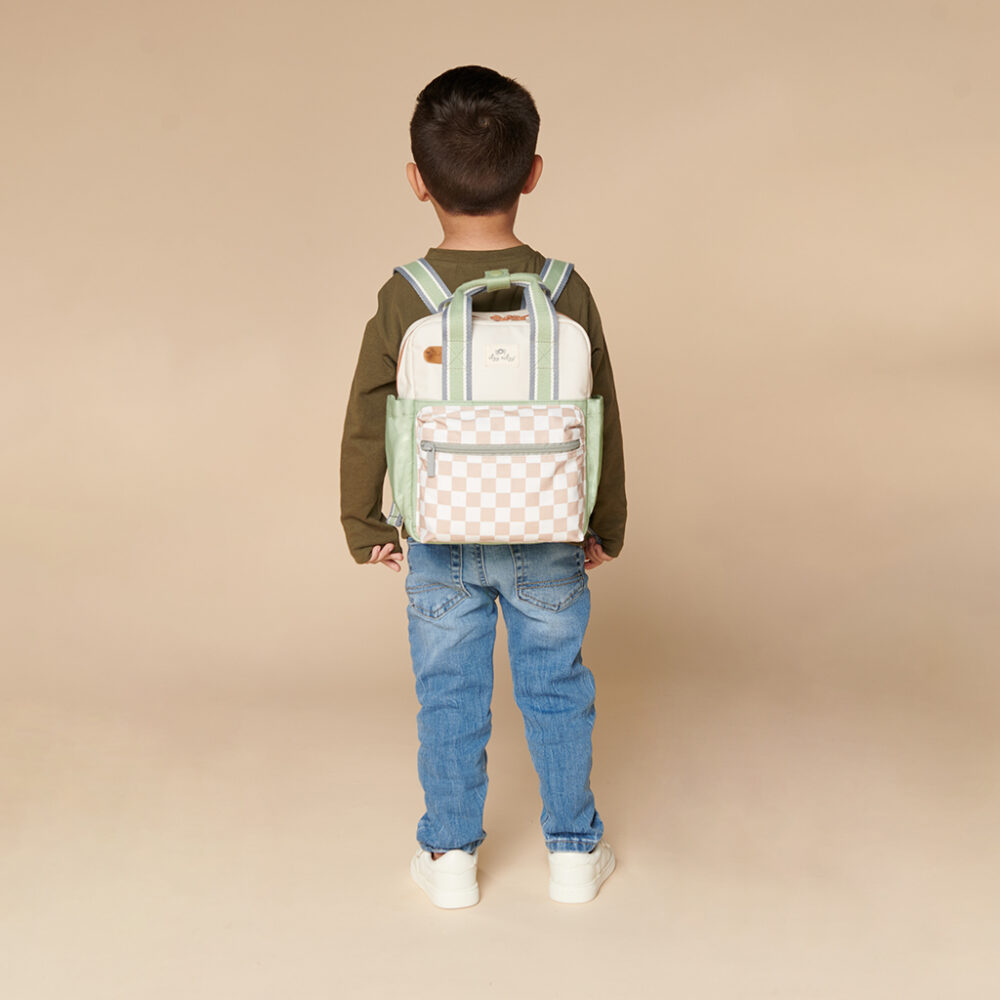 A child with short dark hair, wearing a long-sleeve green shirt, blue jeans, and white shoes, is shown from the back with a colorful backpack. They have an Itzy Ritzy Sweetie Strap Plus Bear Silicone Pacifier Clip attached to their outfit. The background is plain beige.