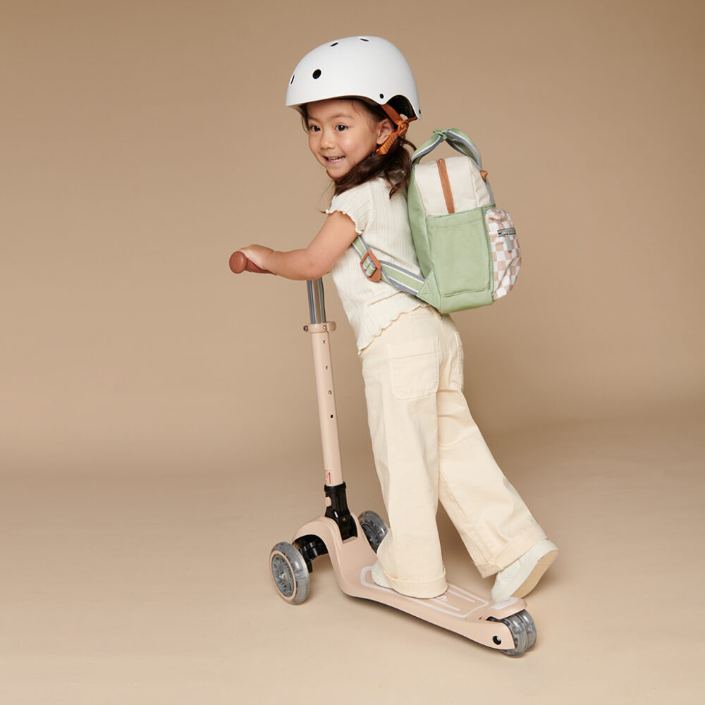 A young child wearing a helmet and backpack rides a three-wheeled scooter on a plain background, with an Itzy Ritzy Sweetie Strap Plus Bear Silicone Pacifier Clip attached to their outfit.