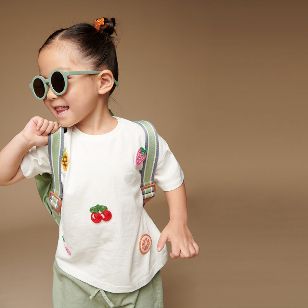 A child with a ponytail and sunglasses smiles while wearing a white T-shirt with fruit designs, green shorts, a backpack, and sporting the Itzy Ritzy Sweetie Strap Plus Bear Silicone Pacifier Clip.