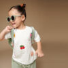 A child with a ponytail and sunglasses smiles while wearing a white T-shirt with fruit designs, green shorts, a backpack, and sporting the Itzy Ritzy Sweetie Strap Plus Bear Silicone Pacifier Clip.