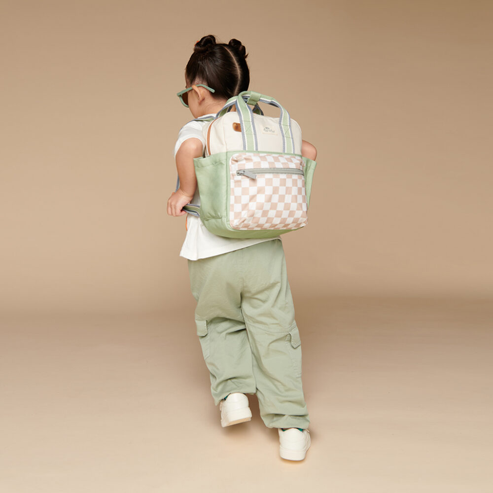 A child with two buns in their hair, dressed in a white T-shirt, mint green pants, and white sneakers, is seen carrying the Itzy Ritzy Sweetie Strap Plus Bear Silicone Pacifier Clip on their mint green and checkered tote backpack over their shoulder as they face away from the camera.