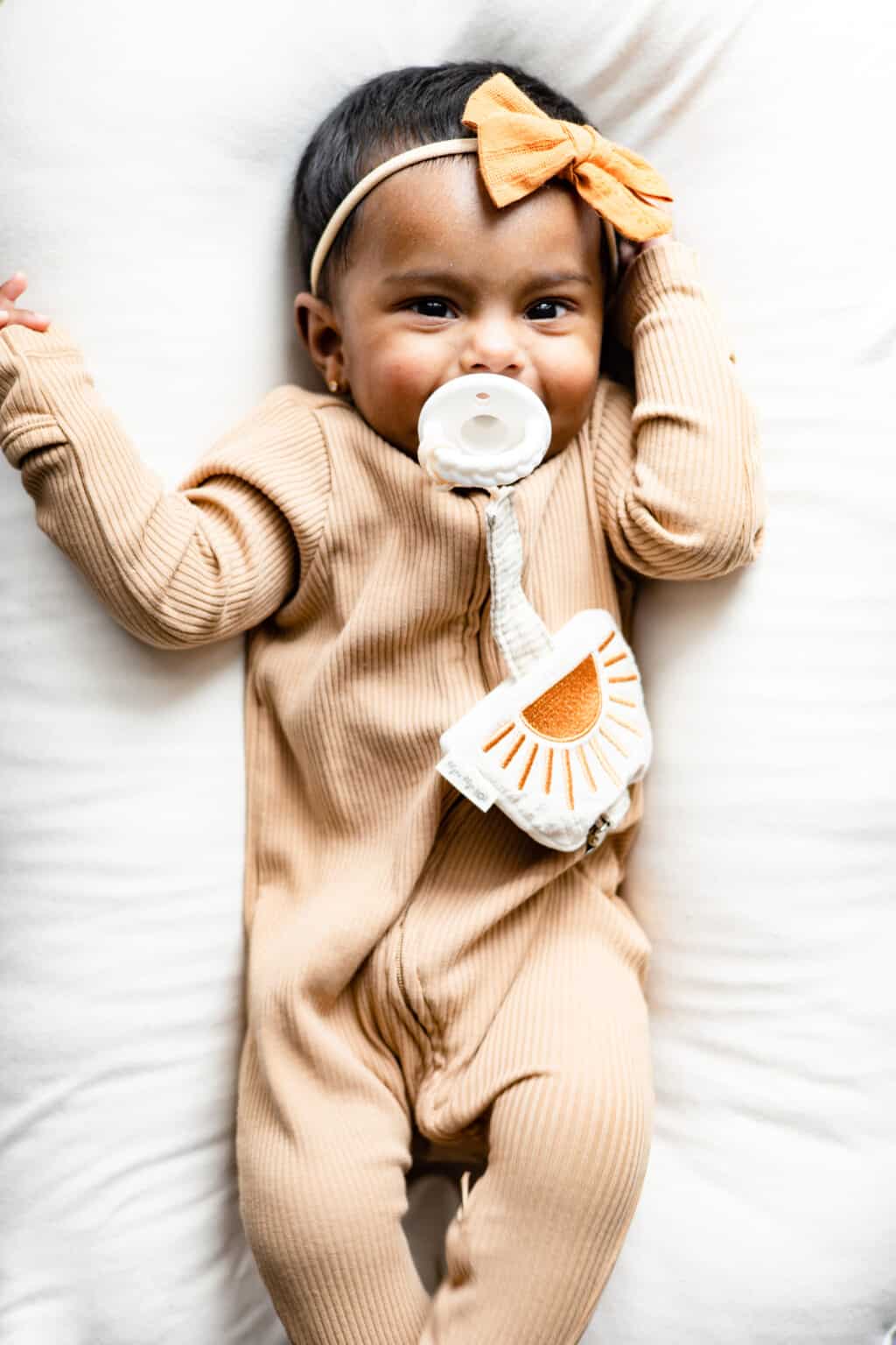 A baby is laying on a bed with an Itzy Ritzy Soothe & Store Pacifier Clip & Storage Pouch in her mouth.