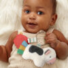 A baby lies on a white furry surface, holding a plush toy with a cow and a red barn teether. The baby wears a beige knitted outfit.