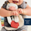 A baby holds a plush cow toy with colorful tags and a red barn-shaped attachment—text reads "SNUGGLY SOFT LOVEY.