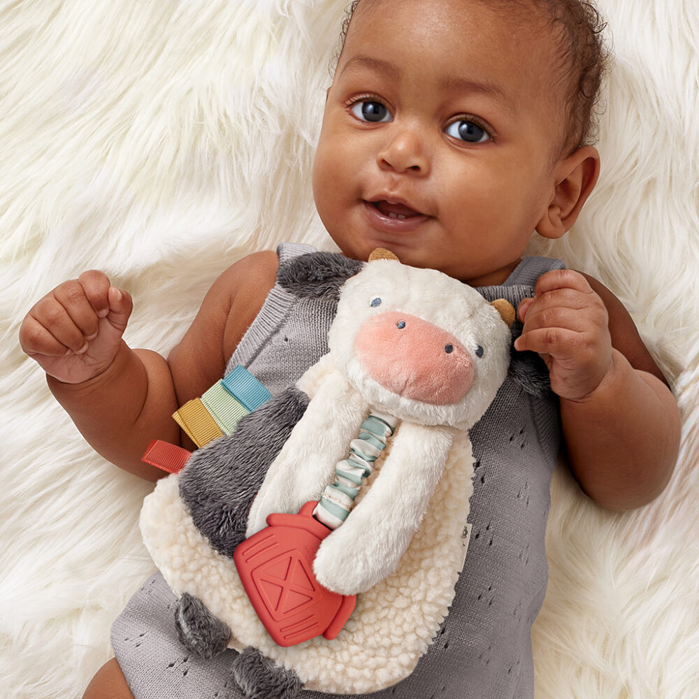 A baby lies on a soft white surface, holding a plush cow toy with colorful tags attached. The baby is wearing a sleeveless grey onesie and looks directly at the camera.