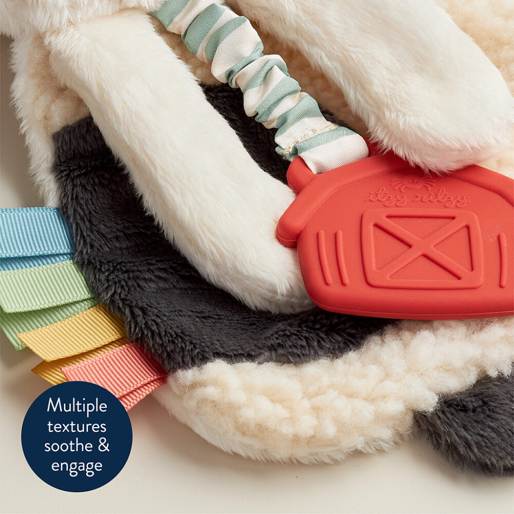 Close-up of a baby sensory toy with various textures including soft fabric, ribbon tags, and a red silicone teether. A tag reads, "Multiple textures soothe & engage.