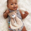 A baby in a gray outfit lies on a fluffy white surface, holding a plush bear toy with a heart-shaped pacifier.