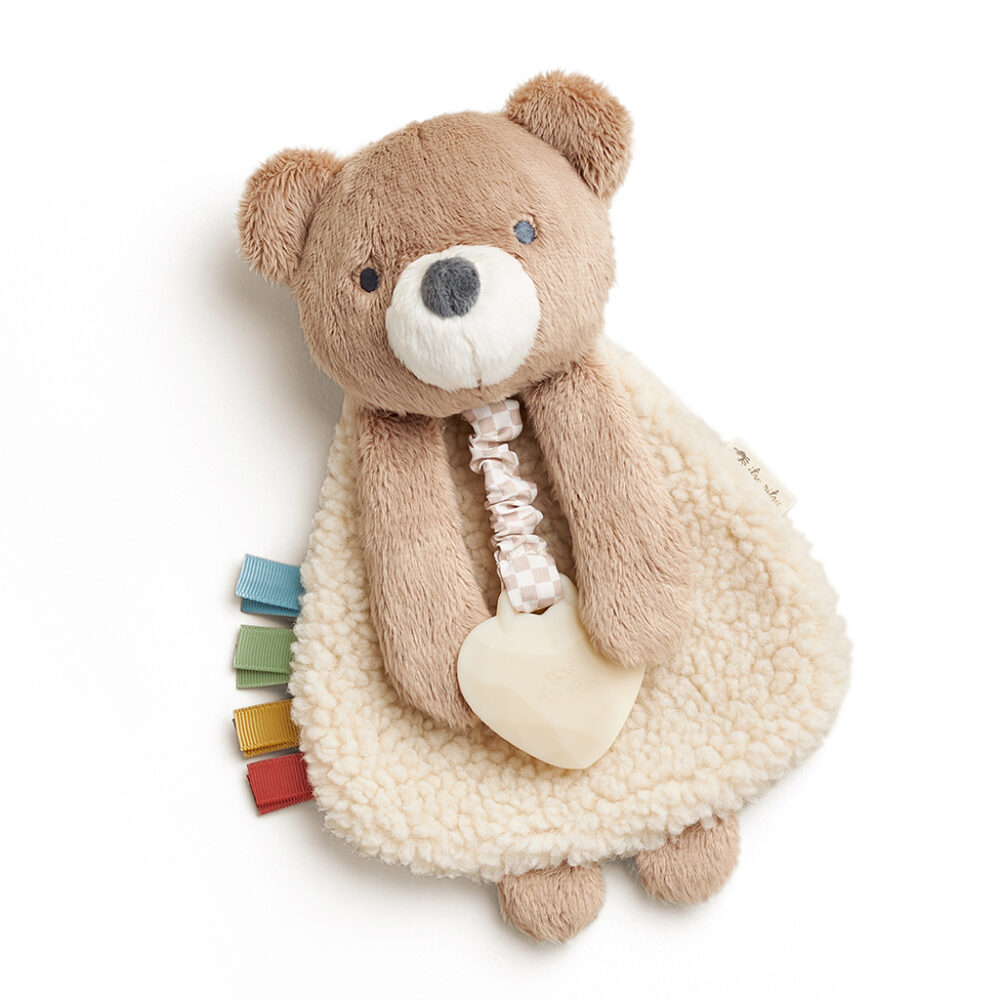 A soft teddy bear plush toy with a white blanket-like body, holding a heart-shaped teething ring. The toy has colorful ribbon tags attached at the side.