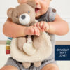 A baby holds a teddy bear lovey with a soft blanket and attached heart-shaped teether. The text reads, "SNUGGLY SOFT LOVEY.