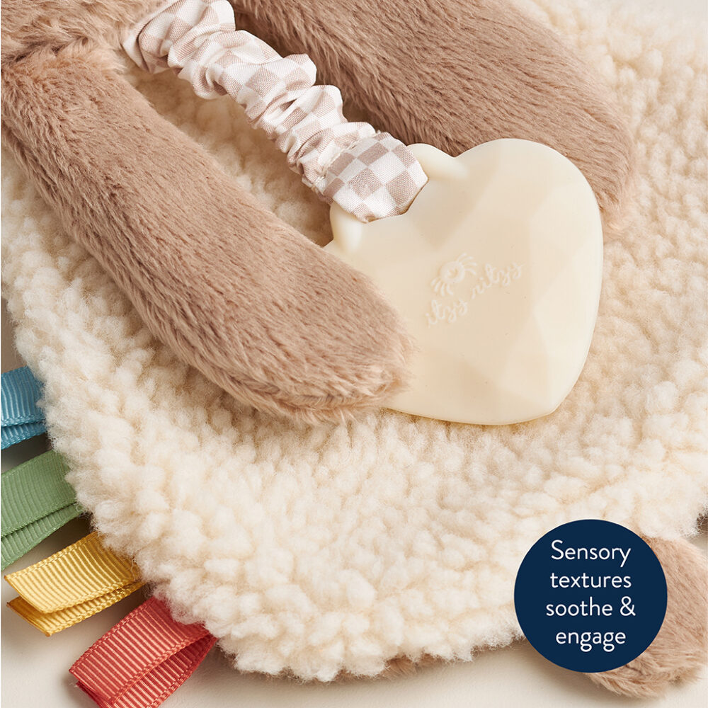 A soft, textured baby toy featuring a plush fabric, a heart-shaped teether, and colorful ribbon tags, with a text bubble that reads “Sensory textures soothe & engage.”.